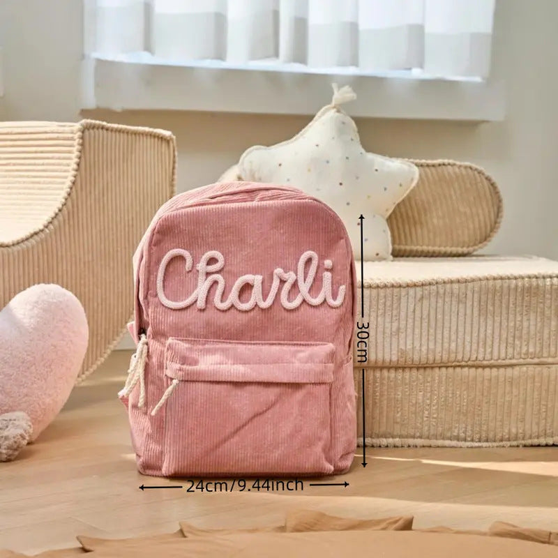 A pink corduroy mini backpack with 'Charli' embroidered on the front, shown with dimension lines indicating 30cm height and 24cm width, set in a cozy room with a bed and pillows.