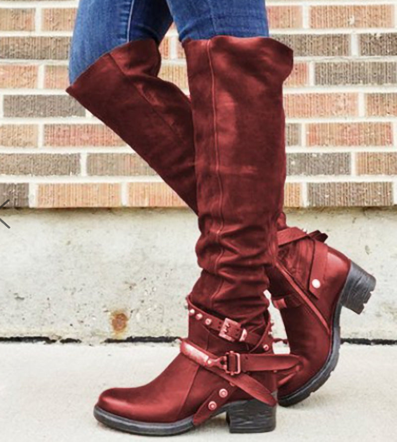 Red knee-high boots with buckles worn by a person against a brick wall.