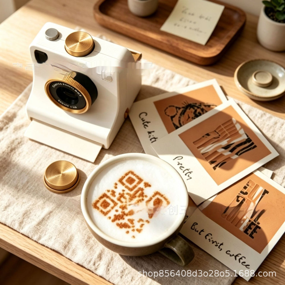 A styled tabletop scene with a white retro camera-like object, a cup of latte with coffee-dust latte art, Polaroid-style cards, and small gold-toned accessories on a beige cloth over a wooden table.