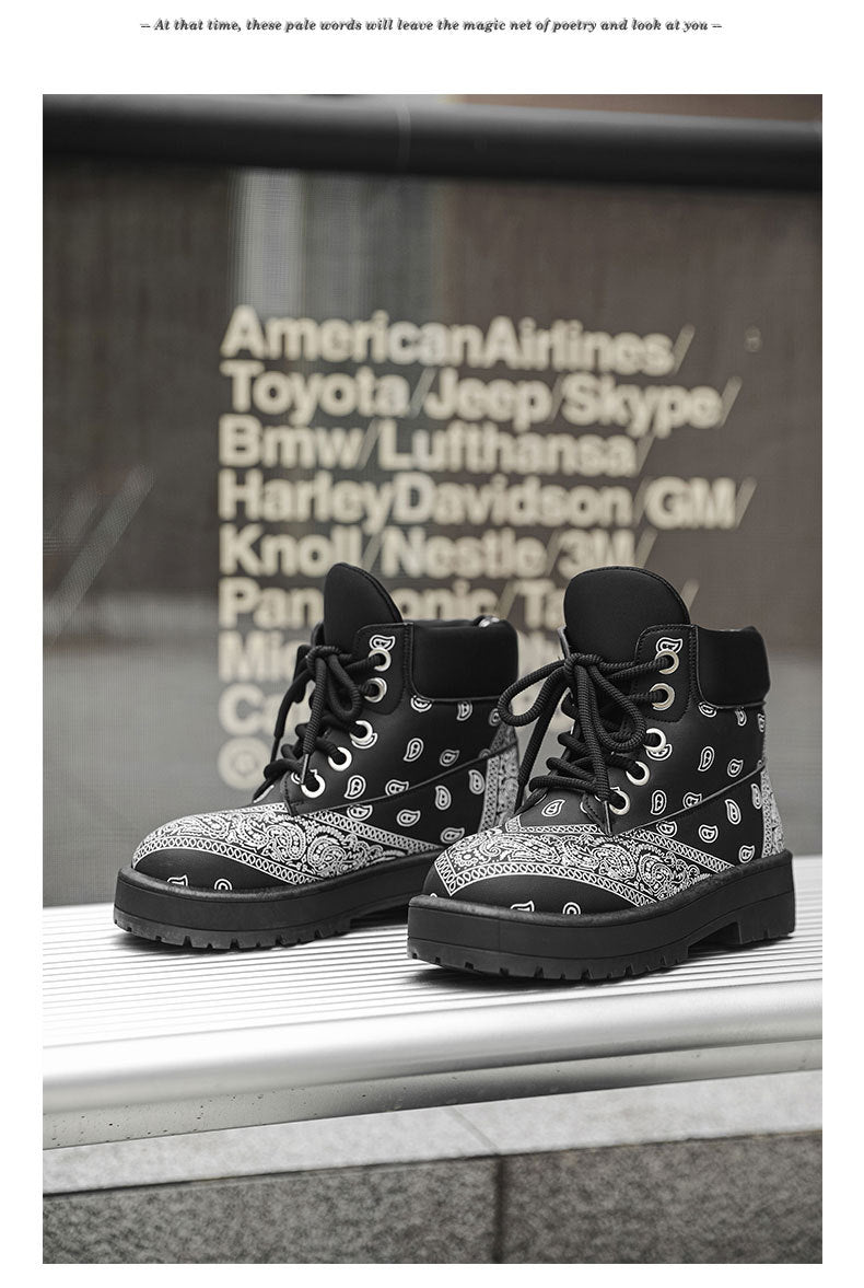 A pair of black and gray lace-up ankle boots with a bandana print pattern, displayed on a white ledge in front of a dark wall with white text.