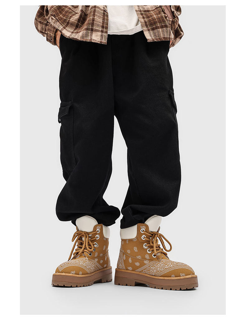 A person wearing loose black cargo jogger pants, a brown plaid shirt, and tan lace-up boots with a subtle pattern, shown from the waist down on a light gray background.