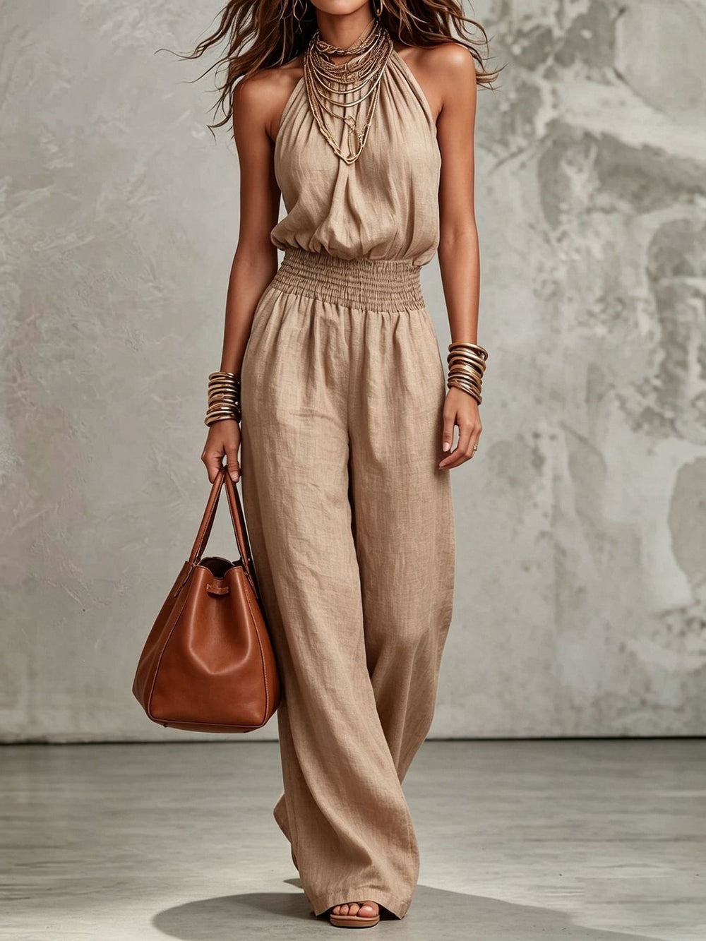 Woman wearing a beige jumpsuit holding a brown handbag against a neutral background