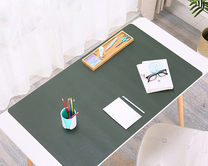 A dark green desk mat laid on a white desk in a bright room, styled with office accessories including a pencil cup, notebook, glasses, and a wooden tool tray.