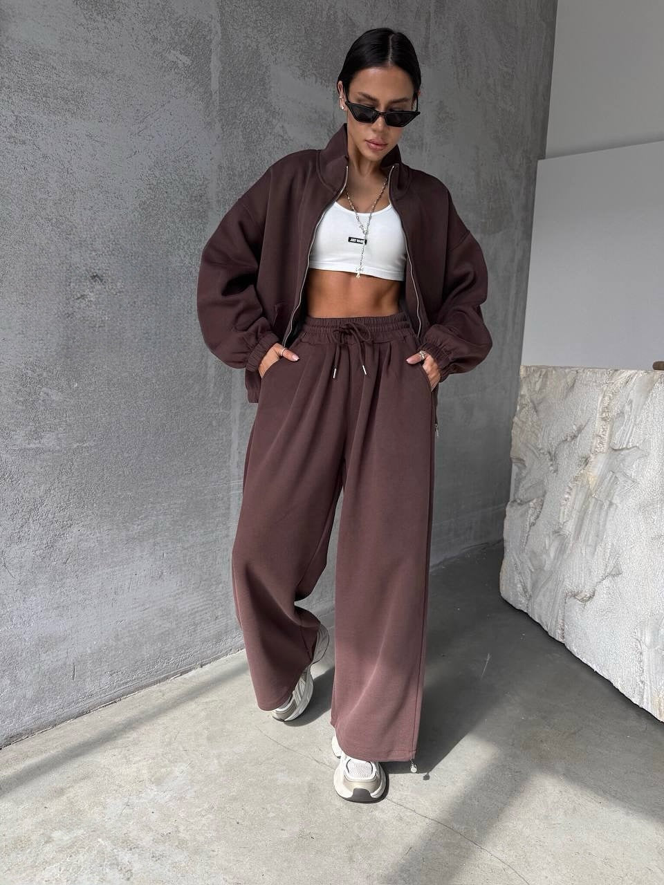 Person wearing a brown tracksuit and white crop top against a gray wall.