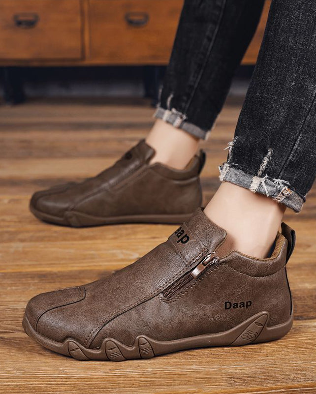 Brown leather shoes with a brand logo worn by a person on a wooden floor.