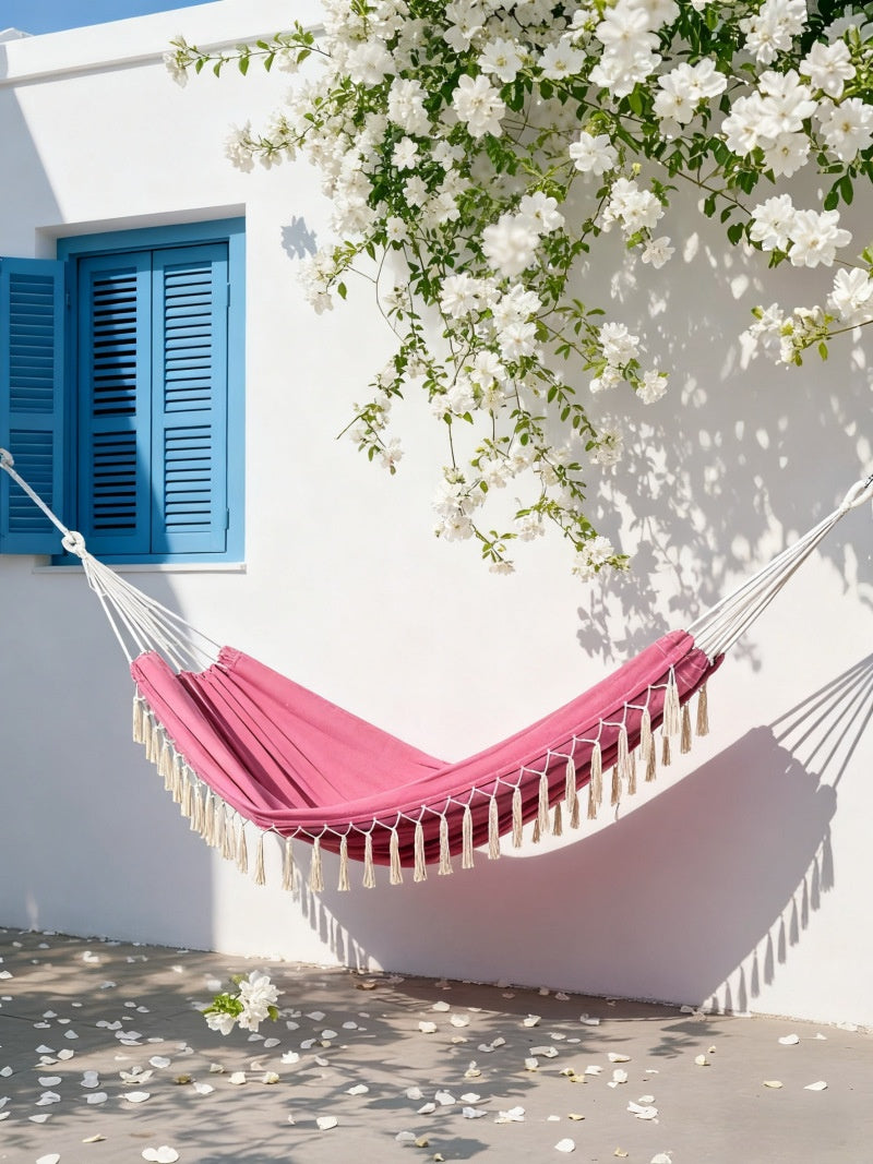 A pink fabric hammock with cream tassels hangs outdoors against a white wall with blue shutters and white flowering vines.