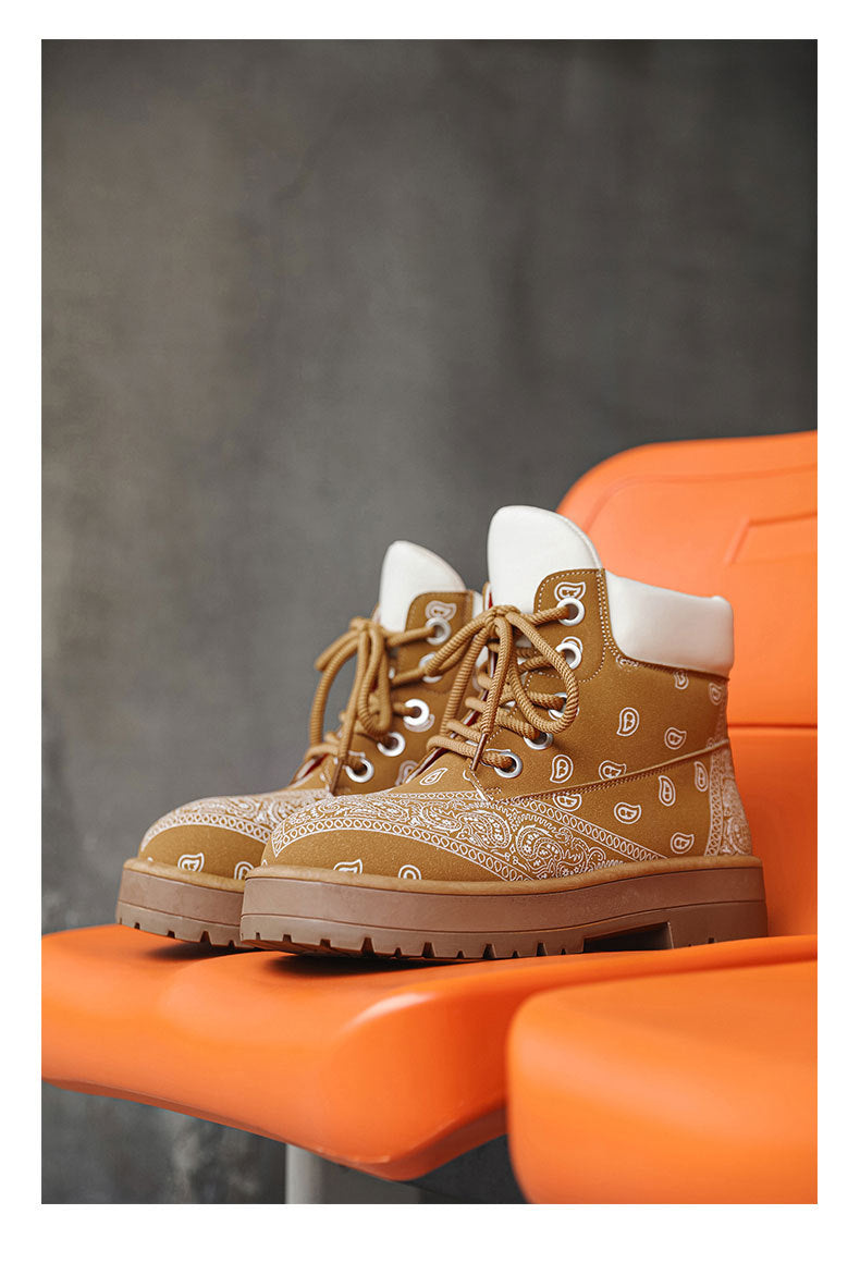 A pair of tan lace-up ankle boots with a white paisley bandana print on the sides, white cuffs, and brown laces, displayed on an orange chair against a dark gray textured background.