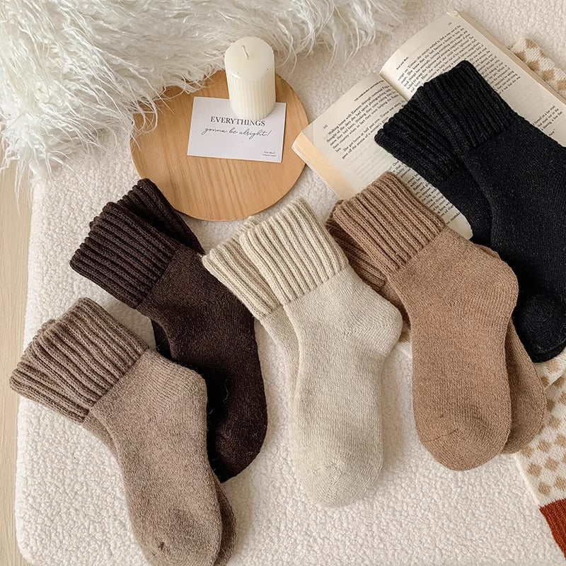 Quattro paia di calze alla caviglia a coste disposte su una superficie morbida e bianca con elementi decorativi tra cui una candela, un libro e un tessuto soffice bianco. Le calze sono disponibili in beige, marrone, crema e nero.