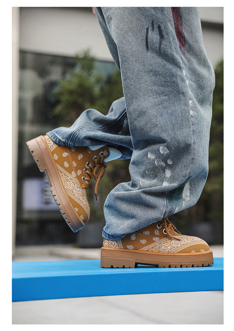 A close-up lifestyle shot of a person wearing light blue distressed jeans and tan bandana-patterned lace-up boots with chunky soles, standing on a bright blue platform outdoors.