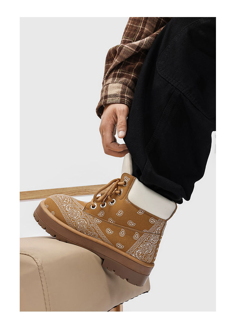 A person wearing a tan bandana-patterned lace-up boot with a white sock, paired with a brown plaid shirt and black pants, sitting on a beige chair against a light gray background.