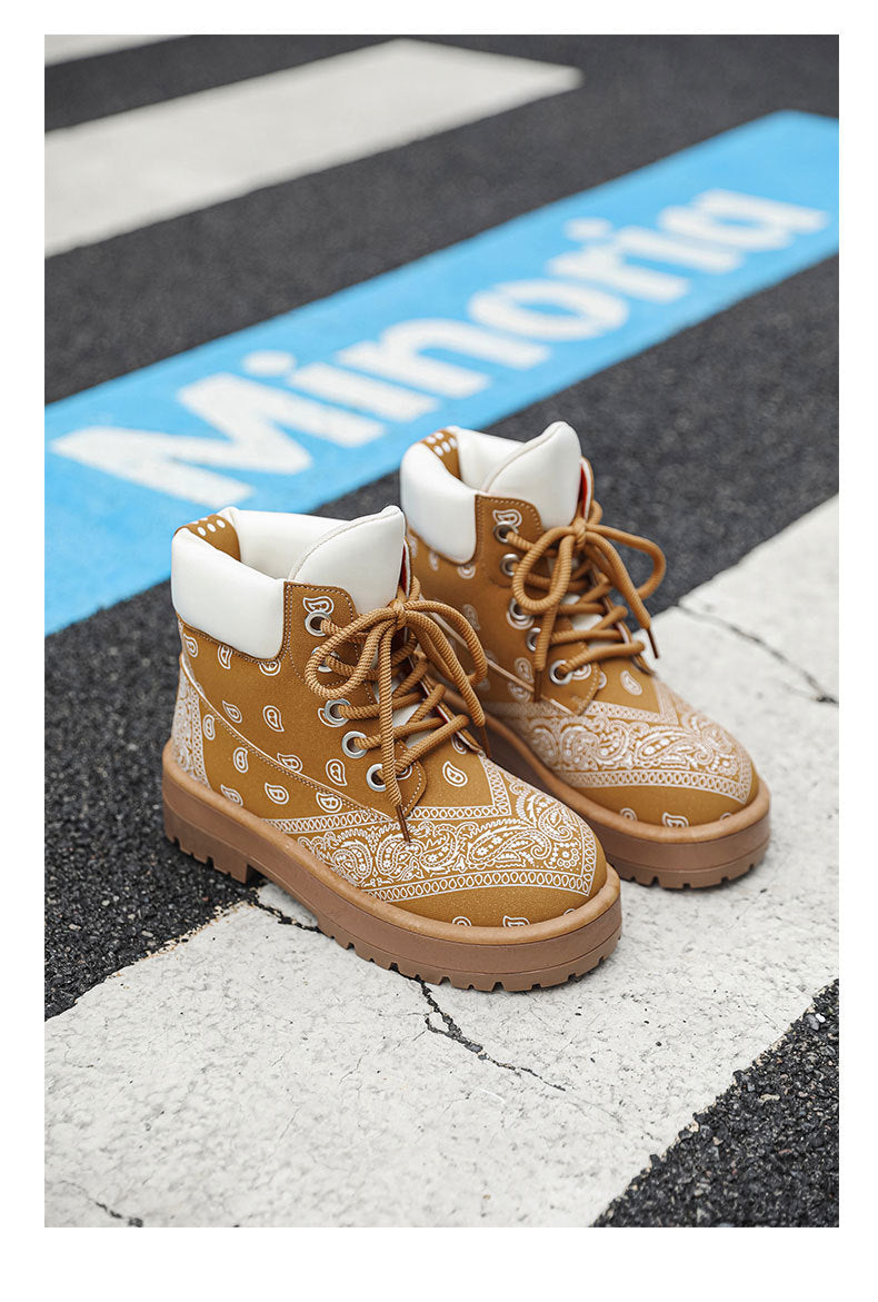 A pair of tan and white bandana-patterned lace-up ankle boots placed on a street crosswalk with a blue sign in the background.