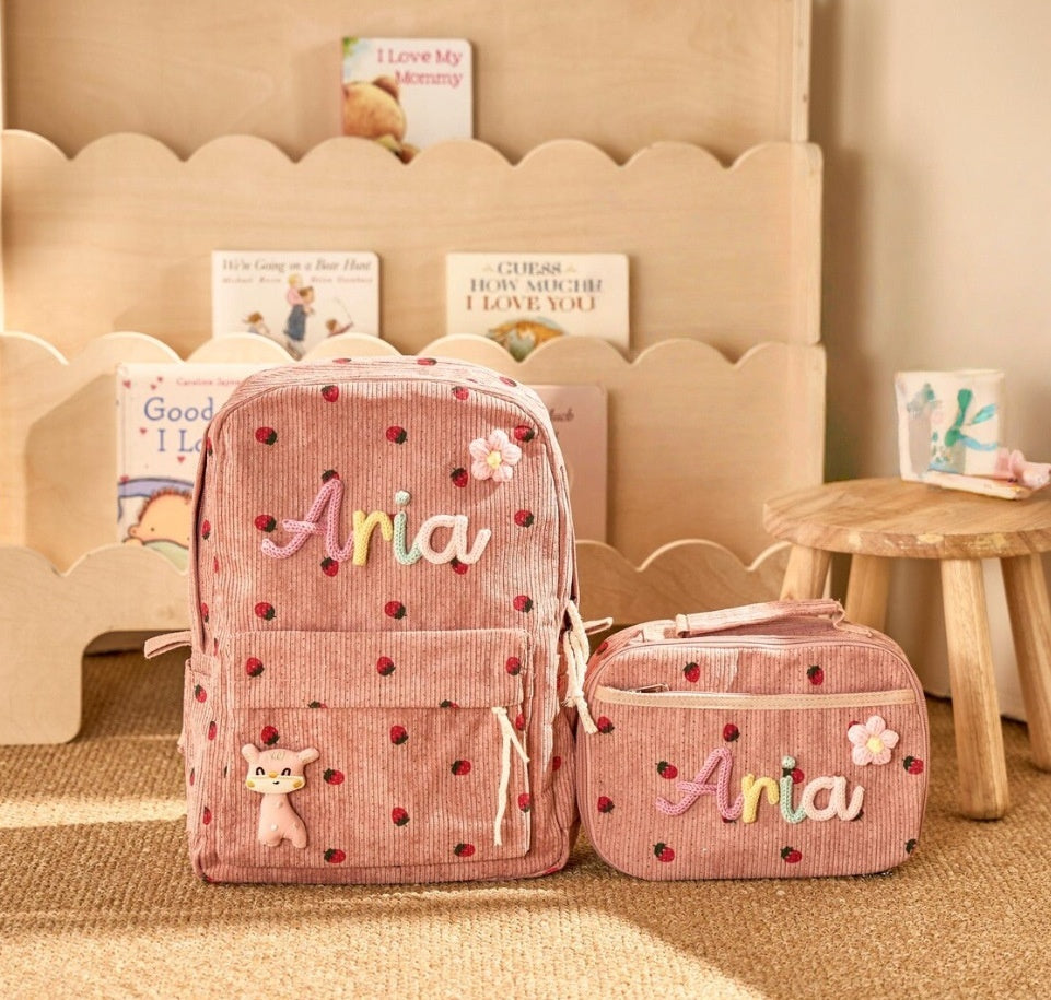 A pink corduroy backpack and matching zip pouch set with embroidered 'Aria' lettering, small strawberry details, and a cute character charm, displayed in a cozy room setting.