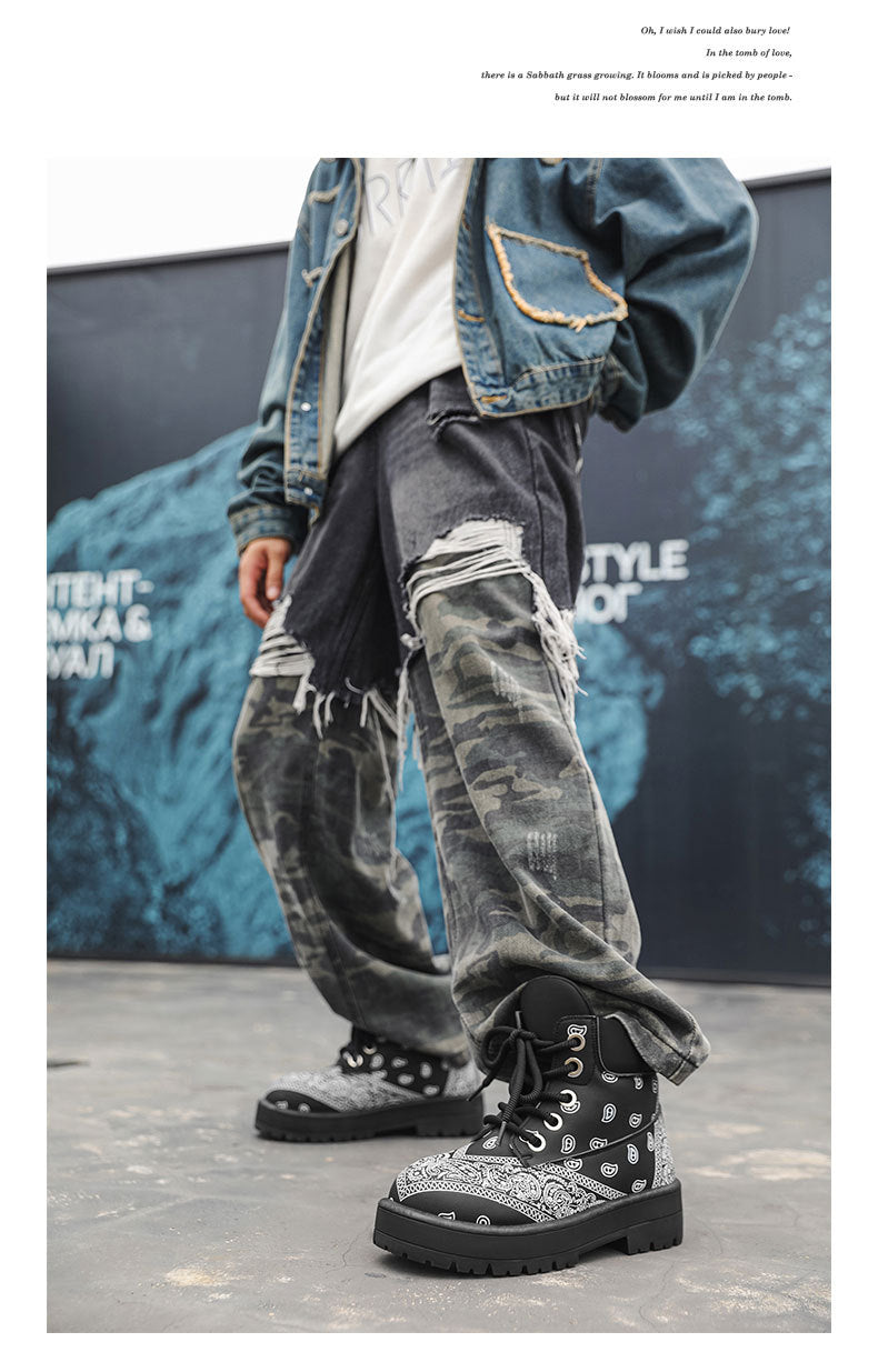 A person wearing distressed gray camouflage jeans, a blue denim jacket, and black high-top boots with white bandana prints and silver studs, standing in front of a dark blue graphic backdrop.