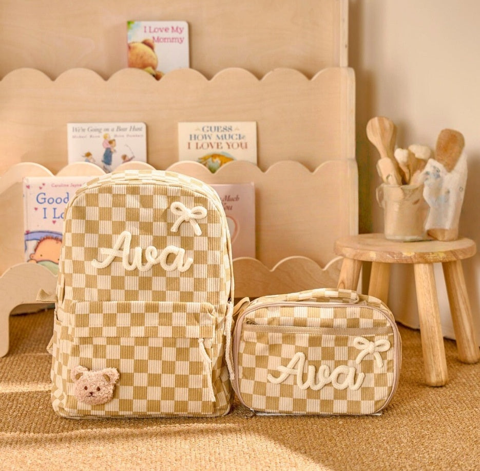 A beige and cream checkered backpack and matching zippered pouch with the name 'Ava' embroidered in white script, styled on a wooden floor in a nursery-like setting.