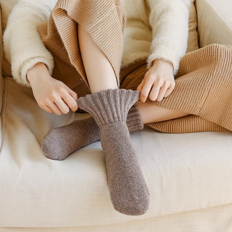 Persona con un maglione beige e pantaloni marroni seduta su un divano beige, che tiene una calza marrone.