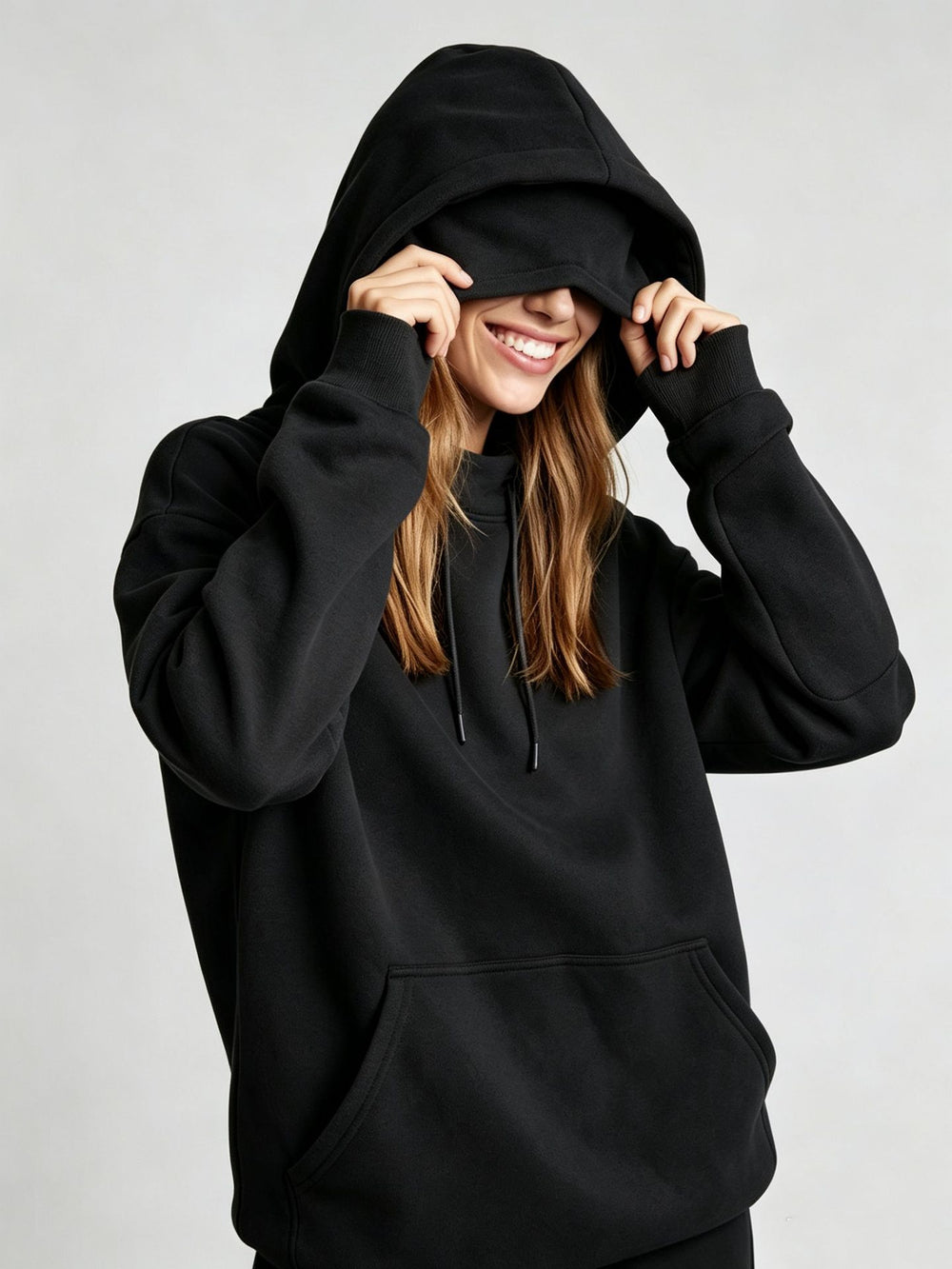 A woman wearing a black pullover hoodie with the hood pulled up over her eyes, smiling and holding the hood in place against a light gray studio background.