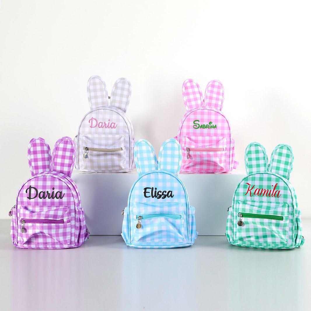 Five personalized bunny backpacks in pastel gingham checks, arranged on white display blocks against a white background. The backpacks feature rabbit ears on the back, a front zip pocket, and custom embroidered names: Daria, Elissa, and Kamila.