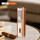 A metallic bronze and brown leather cigar torch lighter stands on a round marble table next to a cigar, with a blurred indoor background. An overlay at the top left reads 'Antique bronze Brown leather'.
