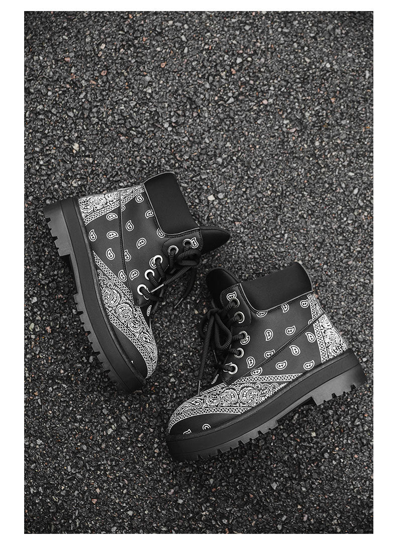 A pair of black high-top lace-up boots with a white bandana print pattern, photographed from a top-down perspective on a dark asphalt surface.