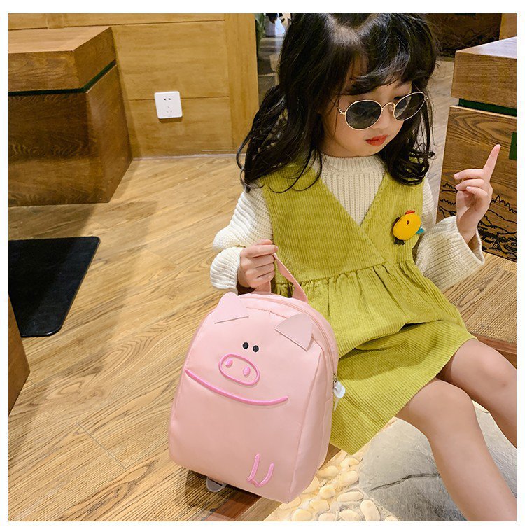 Child wearing a green dress and sunglasses, holding a pink backpack with a pig design.