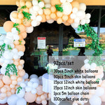 A large balloon arch decoration kit displayed in front of a glass building entrance. The arch is made of clusters of white, peach, and beige balloons with green leafy garland accents.