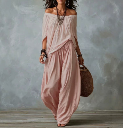 A woman models a dusty pink two-piece bohemian outfit consisting of an off-the-shoulder long-sleeve top and matching loose wide-leg pants, styled with layered necklaces, bracelets, and a woven straw bag against a textured gray studio backdrop.