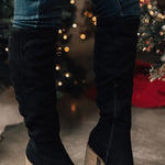 Black suede knee-high boots with wooden block heels worn by a person in front of a decorated Christmas tree.