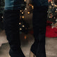 Black suede knee-high boots with wooden block heels worn by a person in front of a decorated Christmas tree.