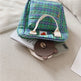 Green plaid bag with a white handle on top of a phone and small round brown object on a beige surface.
