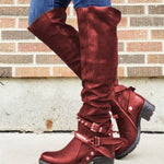 Red knee-high boots with buckles worn by a person against a brick wall.