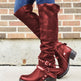 Red knee-high boots with buckles worn by a person against a brick wall.