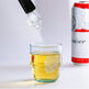 Glass of yellow liquid with a ice cube being added, next to a can of beer on a light gray background