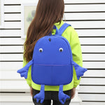 Child wearing a blue backpack shaped like a cartoon character against a white wall.