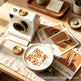 A styled tabletop scene with a white retro camera-like object, a cup of latte with coffee-dust latte art, Polaroid-style cards, and small gold-toned accessories on a beige cloth over a wooden table.