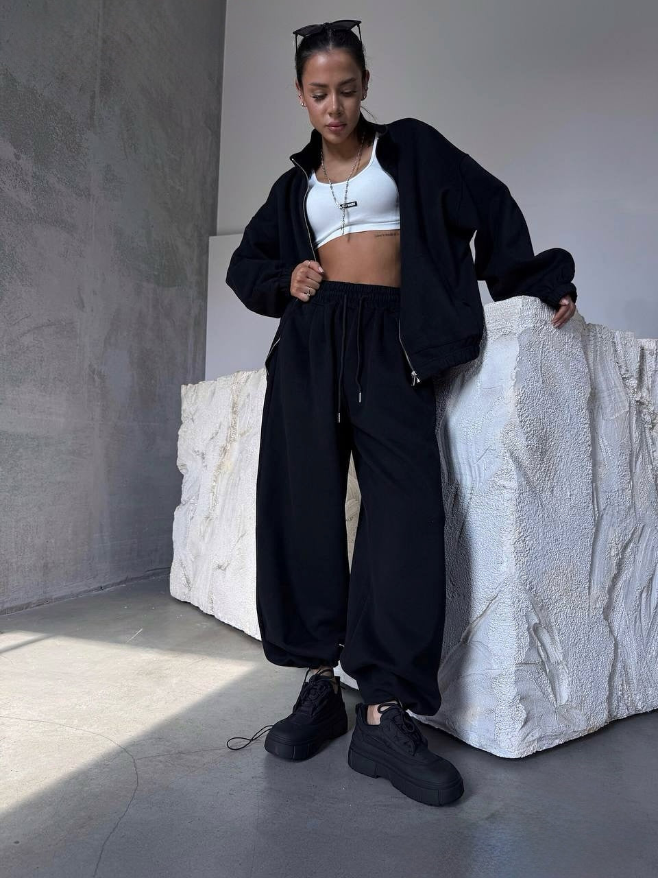 Person wearing a black tracksuit and white crop top in a minimalistic room.