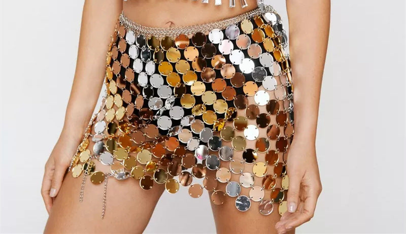 A close-up of a model wearing a short, highly reflective mini skirt covered in large metallic disc embellishments in gold, bronze, silver, and copper tones, with a chain waistband and dangling chains.