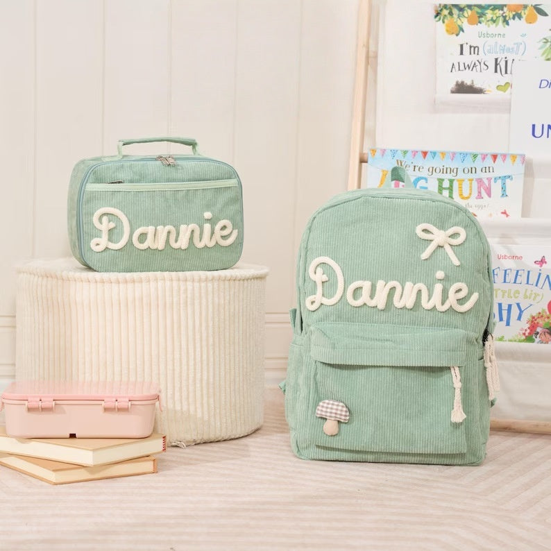 A mint green corduroy backpack and matching lunch bag, both personalized with the name 'Dannie' in white script, displayed in a styled room setting with books and framed art.