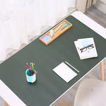 A dark green desk mat laid on a white desk in a bright room, styled with office accessories including a pencil cup, notebook, glasses, and a wooden tool tray.