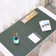 A dark green desk mat laid on a white desk in a bright room, styled with office accessories including a pencil cup, notebook, glasses, and a wooden tool tray.