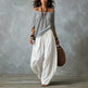 Woman wearing a gray off-shoulder top and white wide-leg pants, holding a woven bag against a gray background.