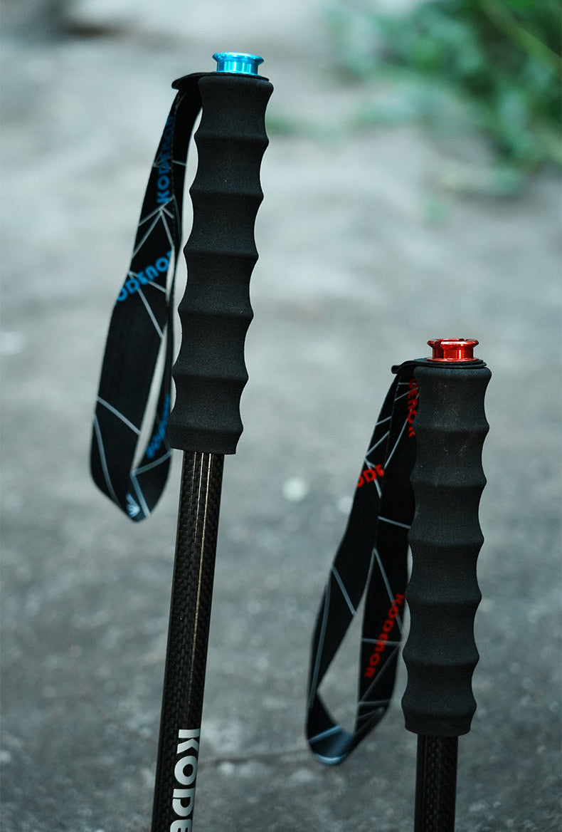 Two hiking poles with textured black grips and wrist straps, one with a blue top cap and one with a red top cap, standing on a gray concrete surface.