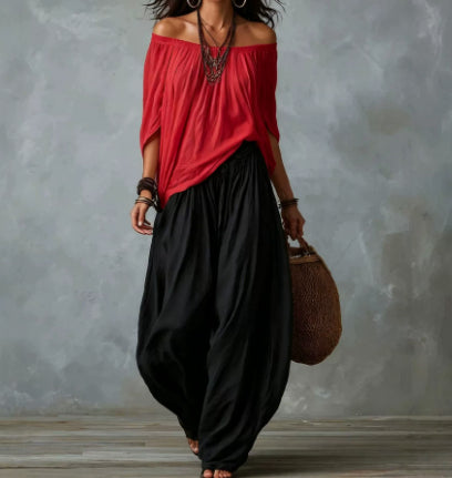 A woman wearing a red off-the-shoulder top and loose black wide-leg pants, holding a brown woven bag against a textured gray wall.