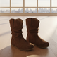A pair of brown suede mid-calf boots with a slouchy, gathered design, displayed on a light wood floor in front of a large window with a city skyline view.