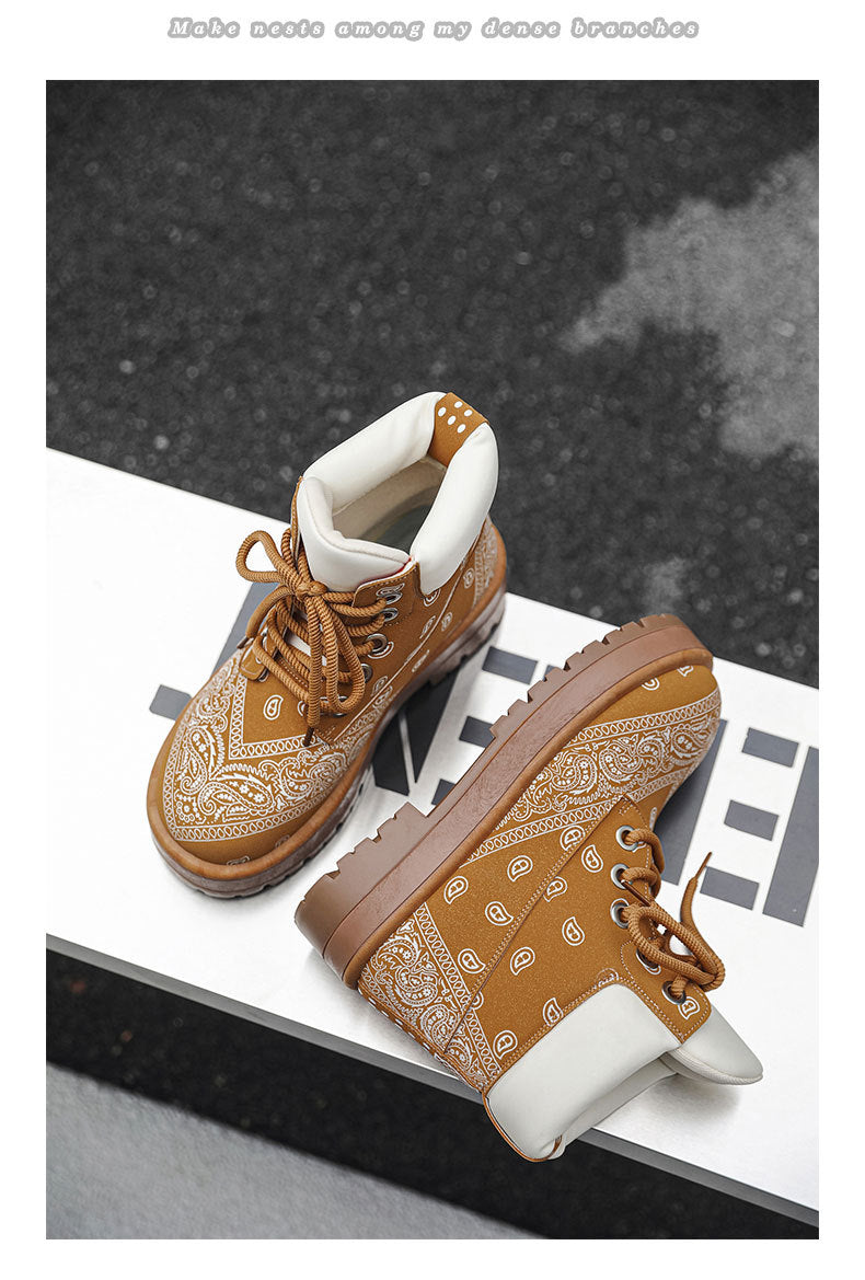 A pair of brown ankle boots with white bandana print overlays, brown laces, and white cuffs, displayed on a white geometric platform against a dark textured background.
