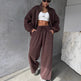 Person wearing a brown tracksuit and white crop top against a gray wall.