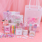 A pink stationery gift set arranged on a pink surface with a pink draped backdrop. The set includes notebooks, pens, a pencil case, sticky notes, stickers, a small card holder, and a large translucent tote bag, all in pastel pink tones with cute cartoon and rainbow designs.