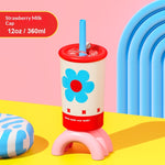 A retro-style 12 oz milk cap tumbler with a blue flower design, red lid, pink stand, and blue straw, set against a pink and yellow background with a blue arch prop.