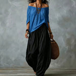 A woman modeling a loose off-the-shoulder blue top paired with voluminous black wide-leg pants, accessorized with layered necklaces, bracelets, hoop earrings, and a woven brown bag, set against a textured gray studio backdrop.