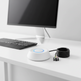 A white circular electronic device with a blue ring around its power button, placed on a white desk next to a coiled black cable and connectors, with a blurred computer monitor and keyboard in the background.