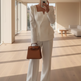 A woman takes a mirror selfie in a minimalist, modern room with beige walls and light wood floors, wearing a white structured blazer and matching wide-leg trousers, holding a brown leather handbag.
